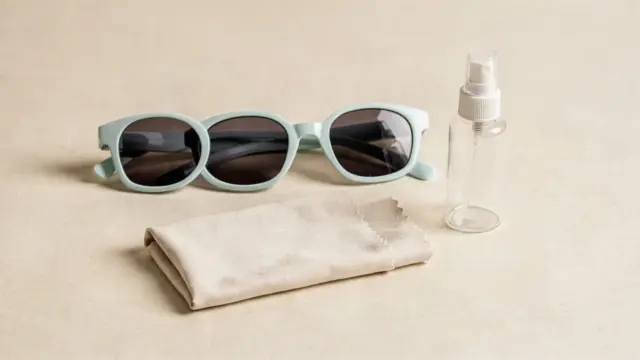 Kindersonnenbrille mit Anti-Beschlag-Beschichtung, Mikrofasertuch und Sprühflasche auf hellem Tisch